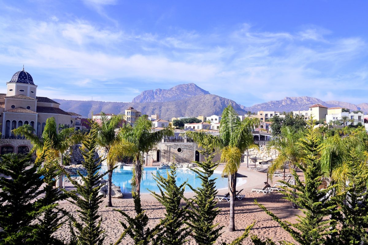 Hotelanlage mit Palmen im Vordergrund und Bergen und blauem Himmel im Hintergrund.