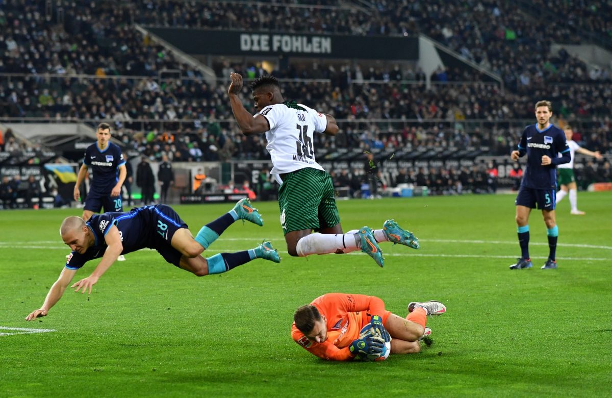 Ein Torwart mit Ball am Boden, über ihm fliegen in verschiedene Richtungen ein Verteidiger (Hertha) und ein Angreifer (Gladbach), um ihm auszuweichen.