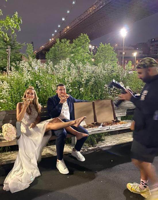 Eine Frau im Hochzeitskleid und ein Mann im Anzug sitzen auf einer Bank und essen Pizza, im Hintergrund eine Brücke.