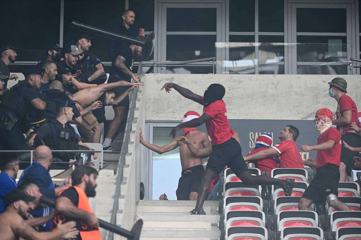 Durch einen Treppenlauf getrennt stehen sich links und rechts Fans beider Teams gegenüber, links vereinzelt auch Polizei. Ein in rot gekleideter Fan wirft einen Gegenstand.