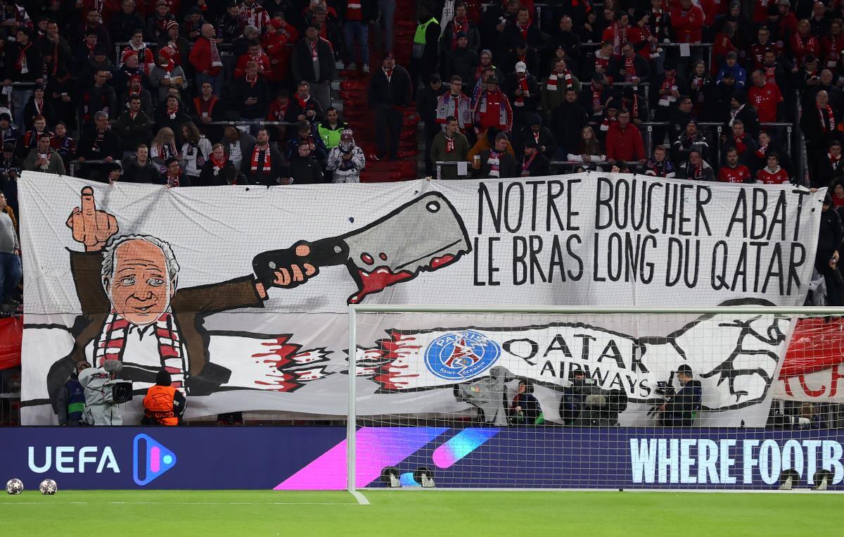 Auf einem Transparent vor der Kurve der Bayern: Uli Hoeneß, ein Mittelfinger, ein Schlachtermesser und auf französisch der Satz: "Unser Metzger schlachtet den langen Arm von Qatar" sowie eine bildliche Darstellung eines zerstörten Arms mit PSG und Qatar-Airways-Logo.
