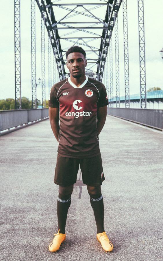 Oladapo Afolayan im FCSP-Heimtrikot der Saison 2023/24. Er steht mit braunem Trikot, Hose und Stutzen auf einer Brücke.