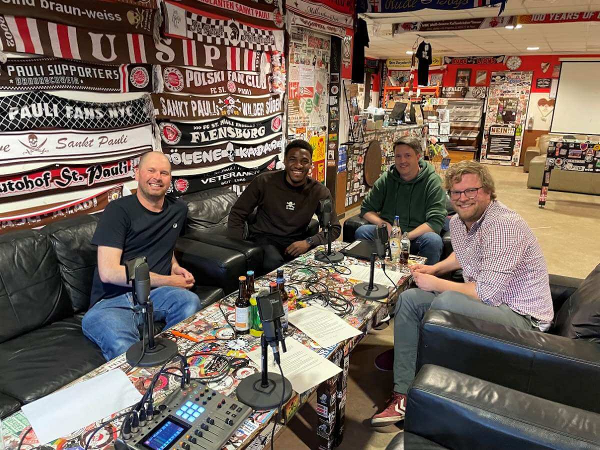 Justus, Dapo, Maik und Tim bei der Podcastaufnahme im Fanladen.
In der Mitte der Tisch mit technischem Equipment, im Hintergrund Fußballschals.