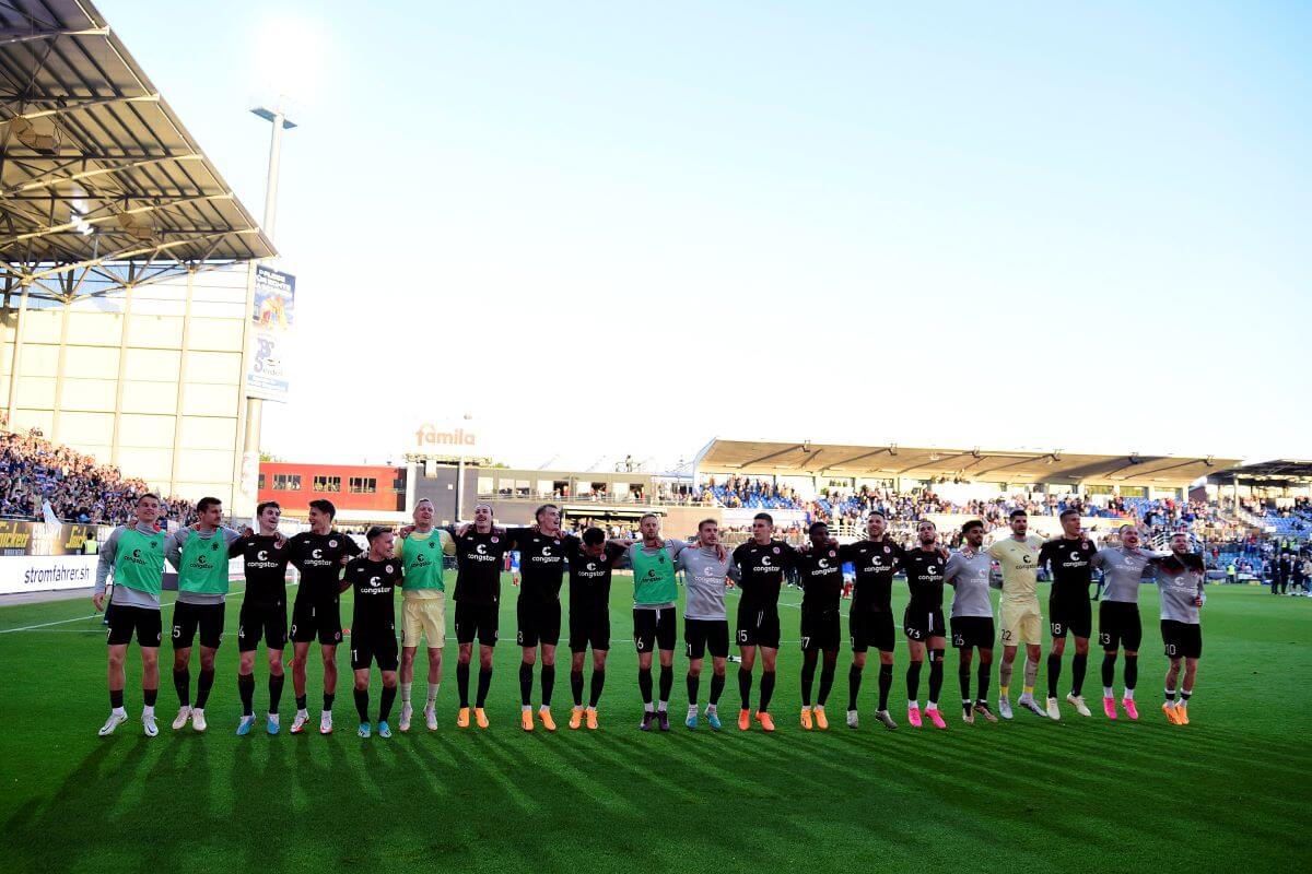 Die Mannschaft feiert nach dem Spiel vor dem Gästeblock in Kiel. Alle haben sich in einer langen Reihe an den Schultern eingehakt.