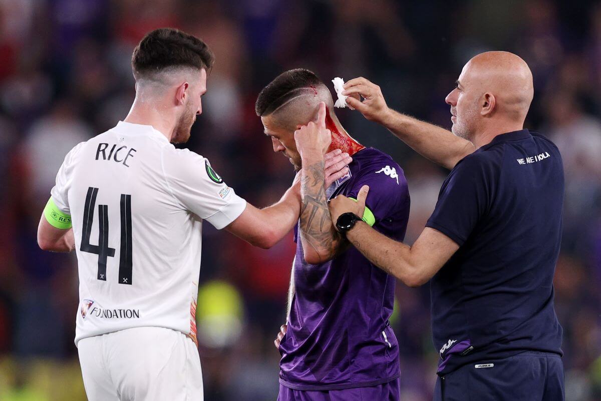 Ein Spieler im weißen Trikot von West Ham (Declan Rice) steht bei Cristiano Biraghi, der von einem Teamoffiziellen gerade an seiner Kopfwunde versorgt wird. Zu sehen ist auch der blutüberströmte Nacken.