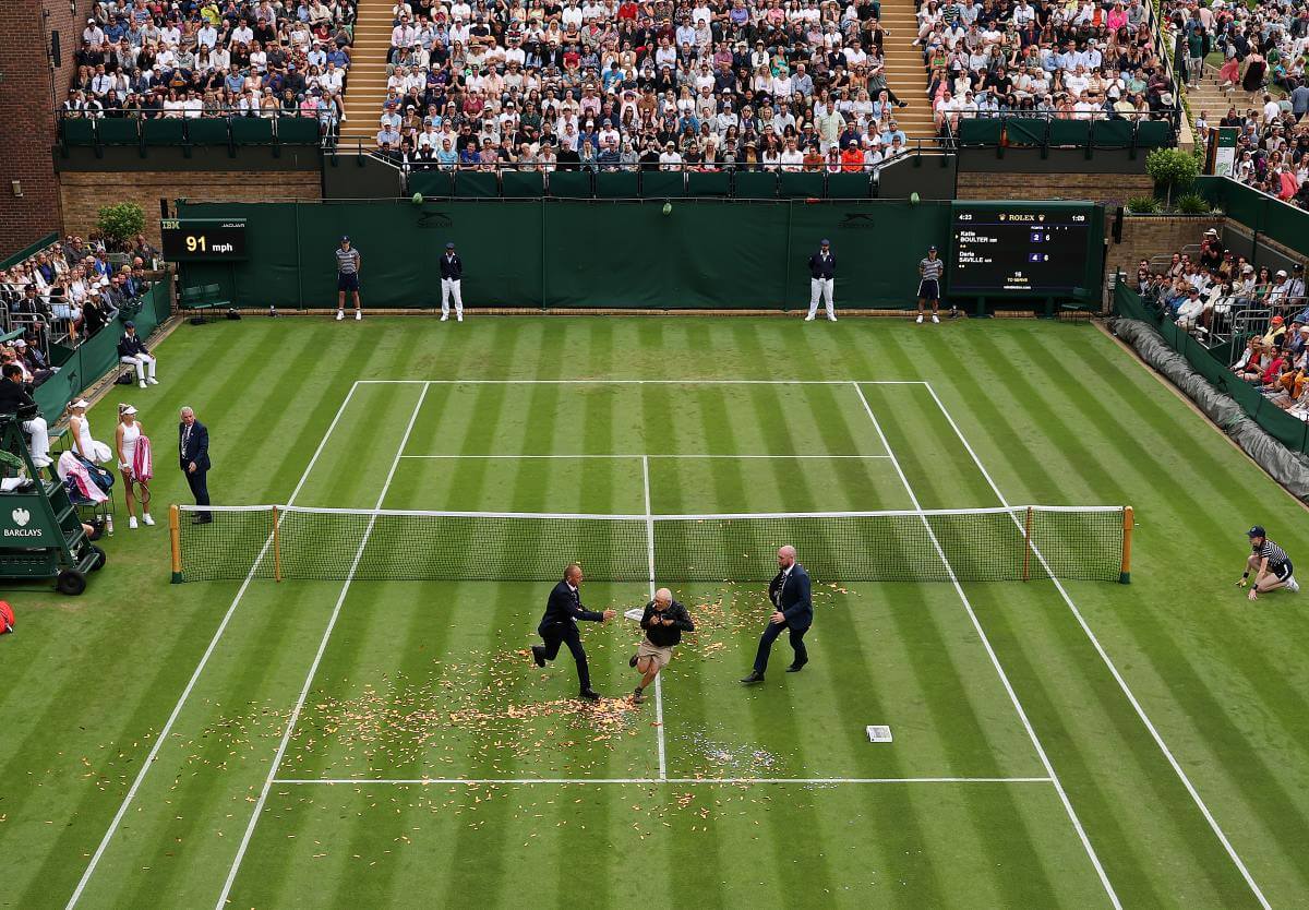 Ein Mann wird auf dem Tennisplatz von zwei Ordnern gejagt, auf dem grünen Rasen liegt sehr viel Glitzer-Konfetti.
