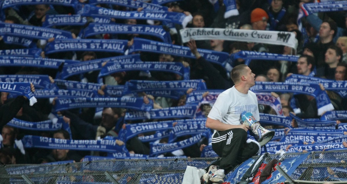 Viele blaue Schals, auf den meisten steht "Scheiss St. Pauli". Davor ein junger Mann auf dem Zaun mit Megaphon.