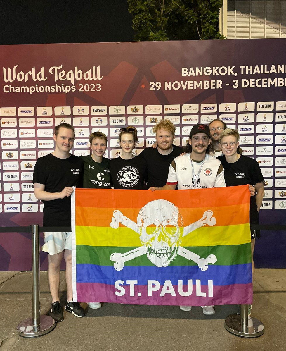 Sieben Spieler*innen mit Regenbogen-Totenkopfflagge vor der Teqball-WM Sponsorenwand.