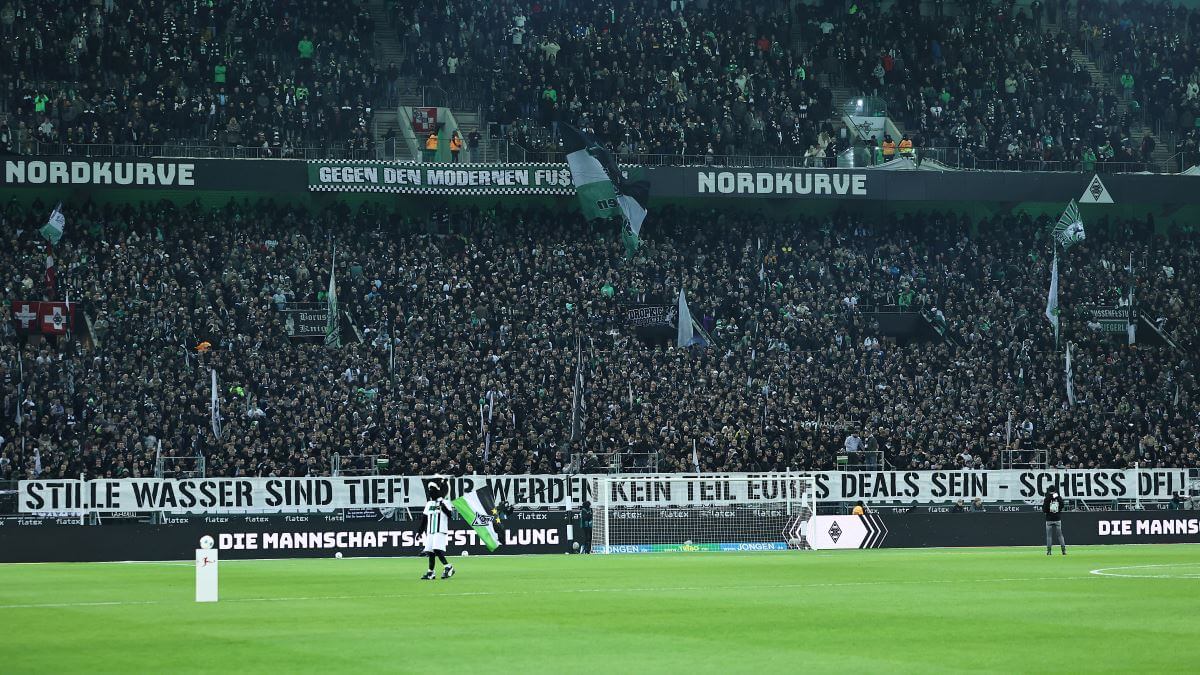 Banner vor der Gladbacher Fankurve vor dem Spiel:
""Stille Wasser sind tief! Wir werden kein Teil Eures Deals sein - Scheiß DFL!"
Vor dem Banner läuft noch das Gladbacher Maskottchen entlang.