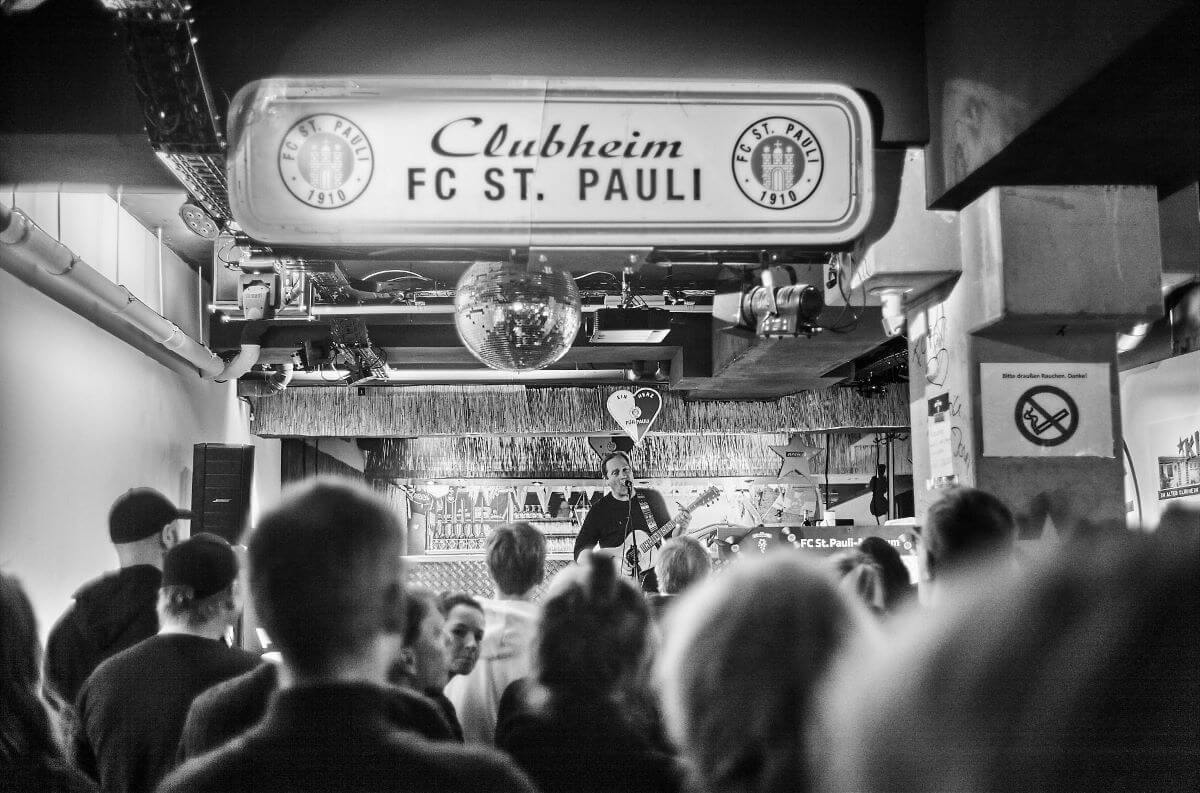 Viele Menschen im Vordergrund, die auf die Bühne schauen, wo Thees Uhlmann mit einer Gitarre steht und singt. Über ihm das alte "Clubheim FC St. Pauli"-Schild.