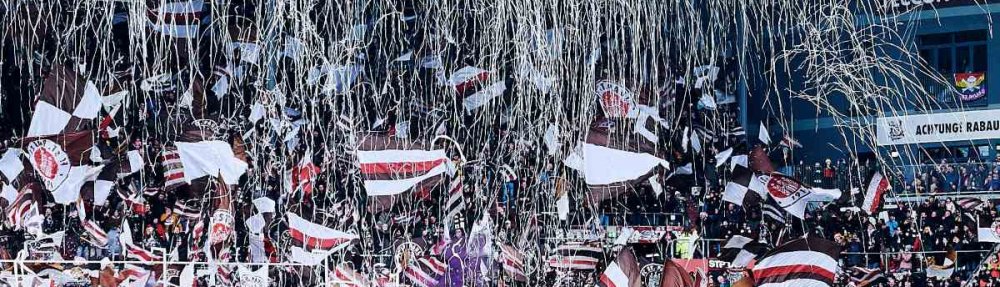 Einlaufen beider Teams beim Spiel FC St. Pauli - Borussia Dortmund vor einer Südkurve am Millerntor, die in Konfetti und Fahnen getaucht ist.
