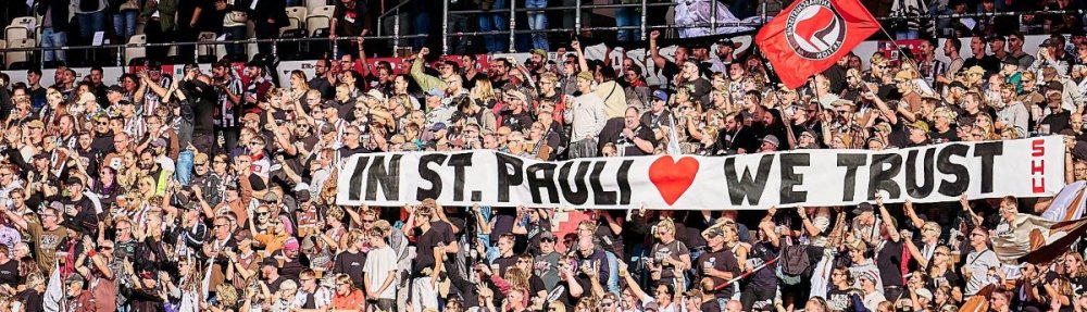 Fans des FC St. Pauli halten auf der Gegengeraden ein Banner mit der Aufschrift "In St. Pauli We Trust" hoch.