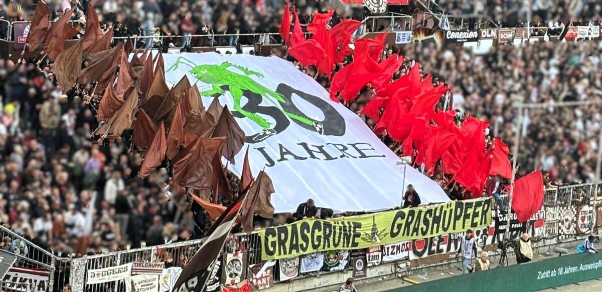 30 Jahre "Grasgrüne Grashüpfer"-Choreo in der Südkurve vor dem Spiel des FC St. Pauli gegen Bayer Leverkusen, 27.09.2025. Zu sehen sind braune und rote Fahnen und in der Mitte das Fanclublogo auf einem großen, weißen Blockbanner.