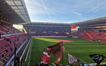 Blick aus dem Gästeblock in die MEWA-Arena von Mainz 05. Im Vordergrund Fahnen, im Hintergrund sich langsam füllende Ränge und ein blauer Himmel.