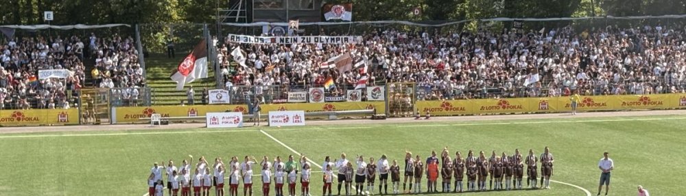 Aufstellung der Teams vom Walddörfer SV und FC St. Pauli 1. Frauen vor dem Hamburger Pokalfinale am 24. Mai 2026 an der Hoheluft. Links der WSV, rechts der FCSP. Im Vordergrund Werbung für das Olympia-Referendum, im Hintergrund eine Protesttapete der FCSP-Fans.