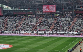 Blick aus dem Gästeblock in Leipzig auf eine sich langsam füllende Heimkurve. Im Vordergrund der Rasen und an der Tribüne der Schriftzug "Leipzig steht zusammen - in jeder noch so dunklen Stunde!"