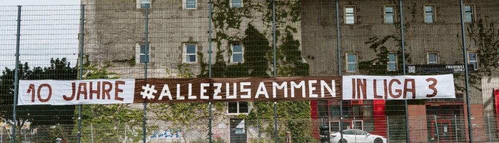 Ein Banner mit dem Schriftzug "10 Jahre #allezusammen in Liga 3" hängt am Zaun der "Feldarena".