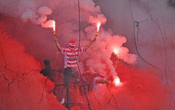 Fan von Energie Cottbus beim Auswärtsspiel bei Hansa Rostock. Der Fan im Trikot ist vermummt, und hat zwei Bengalos in der Hand, um ihn herum ist viel roter Rauch. // (c) Thomas Starke/Bongarts/Getty Images via OneFootball