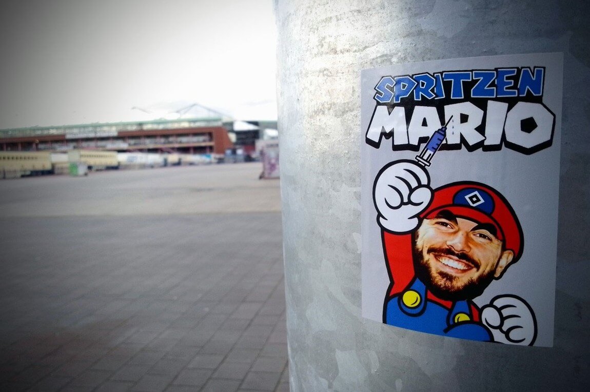 Sticker an einem Pfahl am Heiligengeistfeld, im Hintergrund das Millerntor. Zu sehen ist ein Aufkleber im Super-Mario-Style, beschriftet mit "Spritzen Mario". Der Comicheld trägt eine Raute auf der Mütze, eine Spritze in der hochgereckten Hand und das Konterfei von Mario Vuskovic. // (c) Kiezkieker